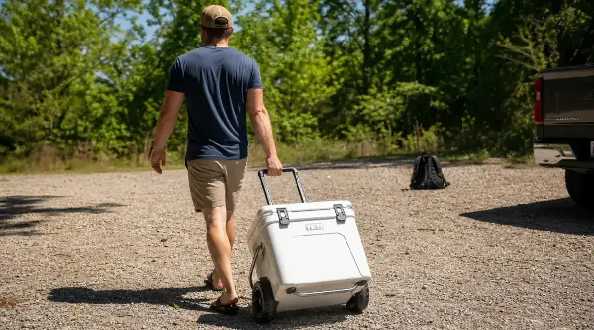 Coleman Classic cooler being rolled across a gravel parking lot toward a campsite, handle extended, wheels visible