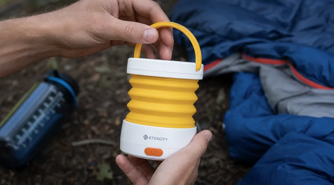 Hand pulling up the top handle of the Etekcity lantern to expand the collapsible body, camping gear in background
